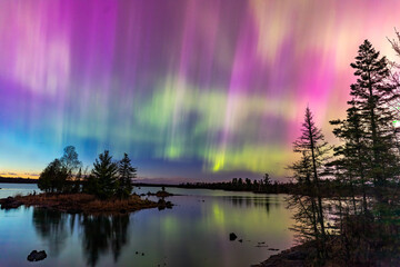 Northern lights erupt over a lake in Minnesota in a dark sky overhead shining rainbow of Aurora light as the sun sets on the horizon