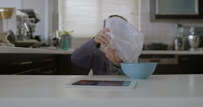 A young boy is seated at a modern kitchen table, focused on a digital tablet with a bowl of cereal in front of him.