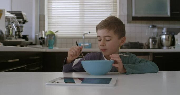 A young boy is seated at a modern kitchen table, focused on a digital tablet with a bowl of cereal in front of him.