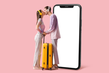 Beautiful young happy couple of tourists in headphones with suitcase kissing on pink background