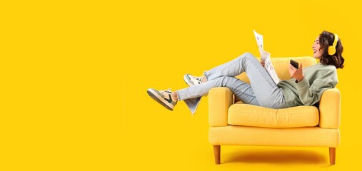 Young woman with newspaper listening to music in armchair on yellow background with space for text