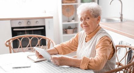 Senior woman with tablet computer writing at table in kitchen