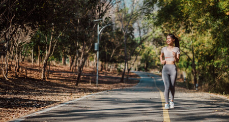 Fototapeta premium A woman is running on a road with trees in the background. She is wearing a pink shirt and grey pants