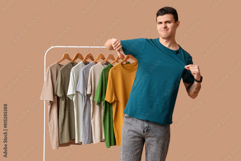 Wall mural Handsome young displeased man showing thumb-down gesture and rack with different t-shirts on brown background