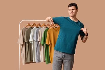 Handsome young displeased man showing thumb-down gesture and rack with different t-shirts on brown...