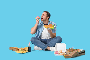 Young man with unhealthy food sitting on blue background. Overeating concept