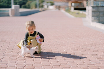 Little girl with a toy in her hand strokes a kitten while squatting on a paved path