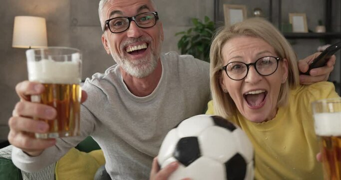 Mature husband and wife watch football match and hold ball and beer