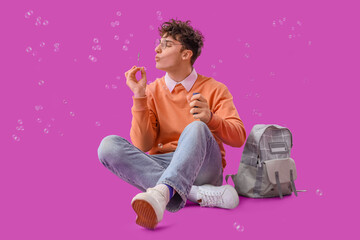 Male student with backpack blowing soap bubbles on purple background