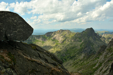 A large stone boulder on the edge of a high cliff overlooking a mountain range on a sunny summer...