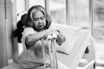 Senior asian woman sitting on bed with crutches at hospital, worried for future or mental health.