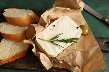 Wooden board with tasty feta cheese, rosemary and bread slices on green background