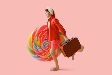 Obraz premium Young man in Santa hat with suitcase and swim mattress running on pink background. Christmas in July