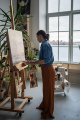 Calm female artist immersed in creative drawing process, painting picture on canvas with oil,...