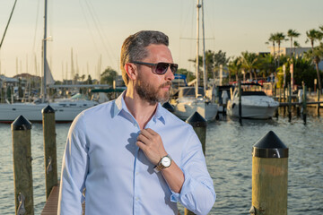 Rich summer business man near the yacht. Portrait of fashion male model outdoor. Rich male posing near yachts. Rich man in urban style. Rich man at yacht club. Man in gulf near luxury yacht ships.