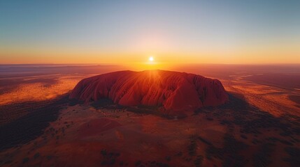 Aerial view of Uluru (Ayers Rock) in Australia, with its massive red sandstone formation rising from the flat desert landscape at sunset.     