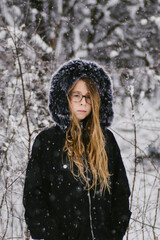 Girl in glasses with long blond hair in coat in snowy forest