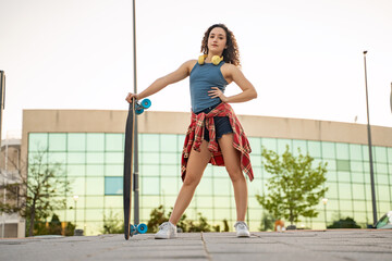 A confident young woman with curly hair holding a skateboard, wearing headphones and a blue tank...