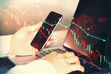 Side view of male hands using smartphone and laptop keyboard at desk with falling red forex chart...