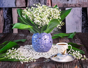 A bouquet of fresh lily of the valley flowers in a blue vase and a white cup of coffee on a dark...