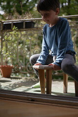 A young boy in a blue shirt sits pensively on a small wooden bench on a porch. The natural greenery...