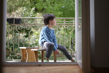 A young boy in a blue shirt sits thoughtfully on a small wooden bench on a porch, looking out at...