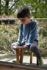 A young boy in a blue shirt, sitting thoughtfully on a small wooden bench outside on a porch. His...