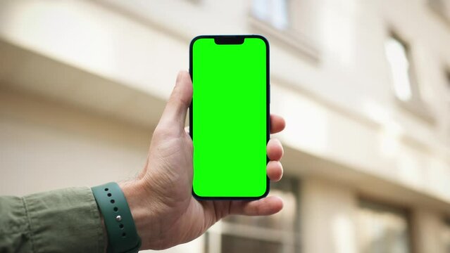 Camera focus on chroma key on smartphone. Person holding mobile device with green screen with one hand. Wonderful yellow building with old ornaments in background. Wearing watch. Sunny weather.