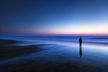 A lone figure stands on a vast empty beach at twilight, reflecting on the horizon, capturing the essence of solitude and tranquility in a serene natural setting