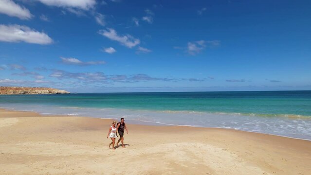 A couple taking a walking hand in hand along a beautiful white sand beach in Australia. The ocean waves can be seen rolling along the shore line in the background as they walk along. Port Willunga