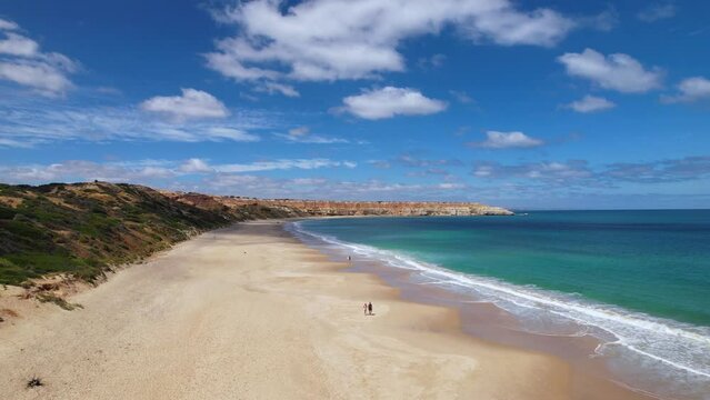 A couple walking along a beautiful beach in South Australia. This is a drone view flying backwards to reveal more of the incredible beach. This is Port Willunga, a white sand beach with a blue ocean.