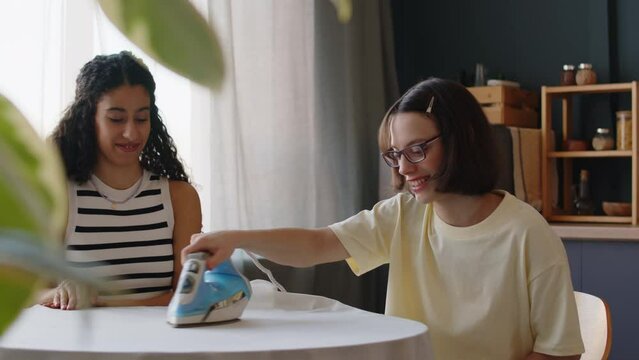 Medium shot of Caucasian girl with cerebral palsy chatting with Biracial female friend while ironing tablecloth on kitchen table in cozy apartment