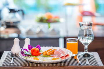 A delicious breakfast of eggs, sausage, ham, and vegetables is served on a white plate with a glass of orange juice and water on the side.