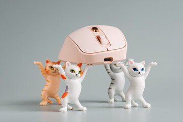 Four funny toy kittens with raised paws carry a pink wireless computer mouse. Light background. A...