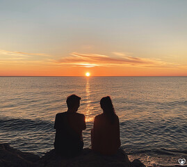 Back view of young couple in silhouette sitting at the beach looking the orange sunset and the horizon over water. Romantic date for lover