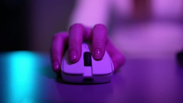 Closeup hand of unrecognizable female freelancer using modern wireless mouse working late evening on computer in dark room with cozy neon light. Woman finger clicking white computer mouse on desk.