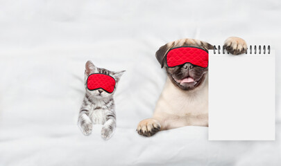 Pug puppy and tiny kitten wearing sleeping masks sleep together on a bed at home. Pug puppy  shows...