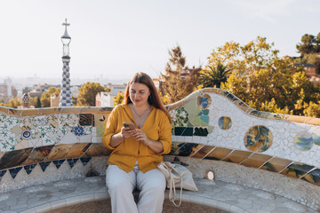 Beautiful female in Gaudi garden, Spain, Barcelona. Young traveling using smart phone outdoors. Concept of travel, tourism and vacation in city