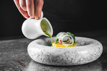 A hand pours green sauce over a gourmet dish on a speckled plate, garnished with basil leaves