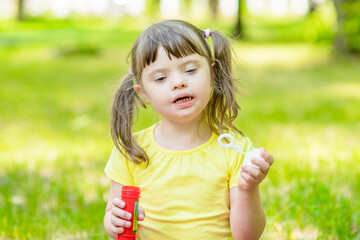 Little girl with syndrome down blows bubbles in a summer park