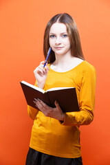 Winsome Blond Girl in Yellow Shirt Posing on Orange Background Studio Portrait as People Lifestyle Concept While Writing Notes in Notebook.