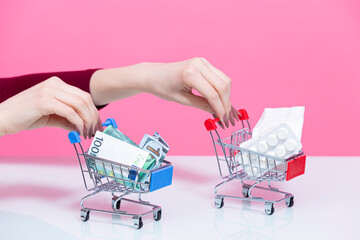 Closeup of Female Hand Holding Mockup Small Tiny Shopping Carts Trolley With Bunch of Medicines and Currency Banknotes On Pink Rose Background.