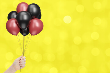 Female hand holds bunch of red and black balloons on blurred yellow background. Empty space for text