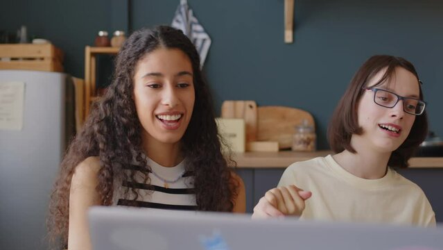 Waist up of happy Caucasian girl with cerebral palsy with Biracial female best friend holding hands and singing while sitting together at table in cozy apartment