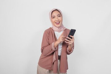 Naklejka premium Excited Asian Muslim woman in hijab pointing to the smartphone she holds over isolated white background.
