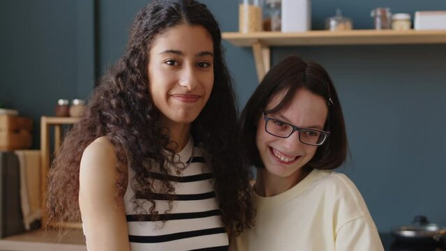 Waist up portrait of cheerful Caucasian teenage girl with cerebral palsy posing for camera with Biracial female best friend at home