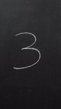 Hand writes the number 3 with white chalk on a black board. Vertical video