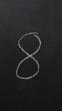 Hand writes the number 8 with white chalk on a black board. Vertical video