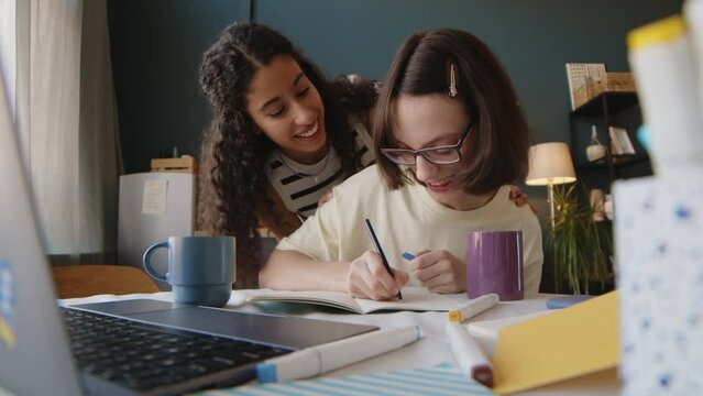 Stab chest up shot of Caucasian girl with cerebral palsy sitting at desk in cozy room and drawing while her Biracial female friend speaking with her and helping