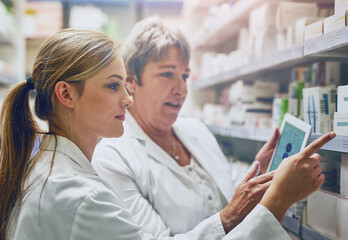 Pharmacist, women and together with tablet by shelf for stock, check and inventory with pointing in shop. People, mentor and intern with digital touchscreen, app and inspection for drugs at store